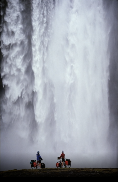 Two bicyclists stand in front of a large waterfall, photo by John Greengo