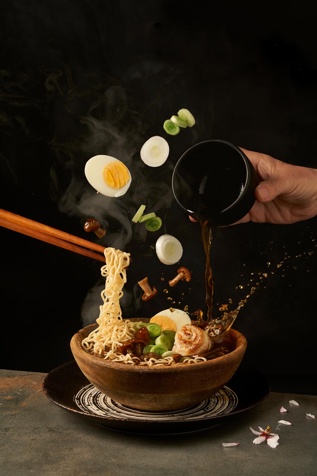 Cook holding noodles with chopsticks and adding miso broth into ramen while eggs, onions, and mushrooms fall into bowl