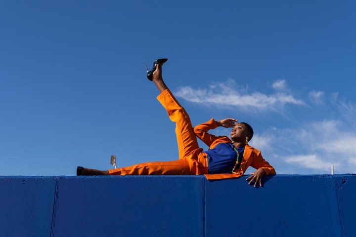 Low angle view of African American female model in orange suit and high heels striking a pose on blue wall