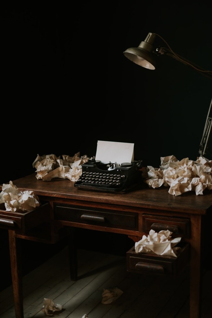 Home office with wooden table and vintage typewriter near old lamp surrounded by crumpled white papers